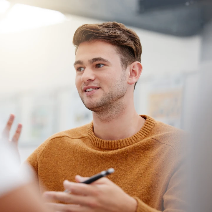 a young man holds a pen and gestures while in a meeting